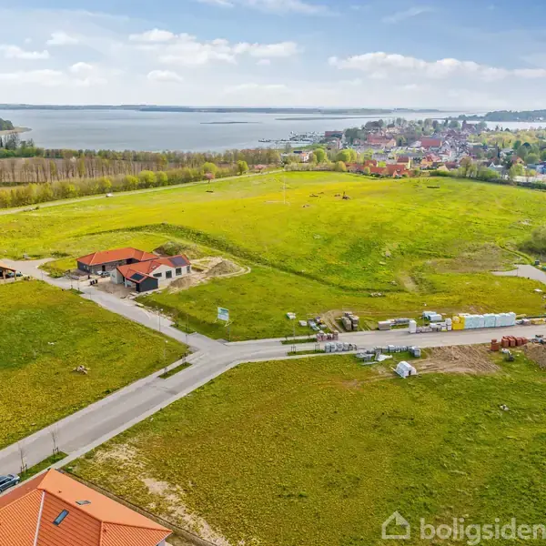 Landskab med græsmarker, flere nybyggede huse med røde tage og en vej. Udsigt til en by og en stor sø i baggrunden under en delvist skyet himmel.