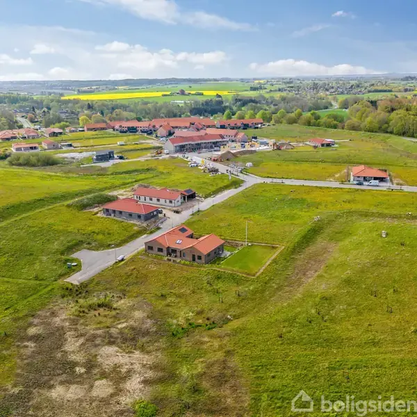 Flere huse med røde tage ligger spredt i et grønt landskab. Veje forbinder bygningerne, omgivet af åbne marker og nogle træer i baggrunden under en klar himmel.