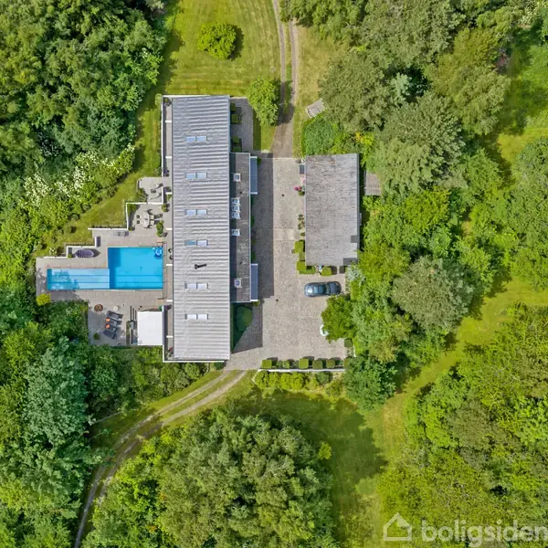 Rektangulært hus med fladt tag, omgivet af frodig grøn have, har en terrasse med pool til venstre. Indkørsel og parkeringsområde set ovenfra, beliggende i en skovklædt landskab.