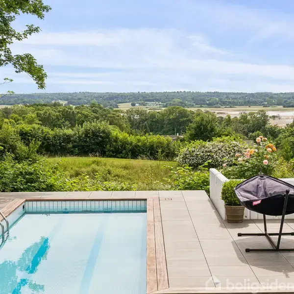 En swimmingpool og terrasse med udsigt over en frodig dal med grønne træer. Parasol og planter pynter terrassen, mens en åben himmel strækker sig i baggrunden.