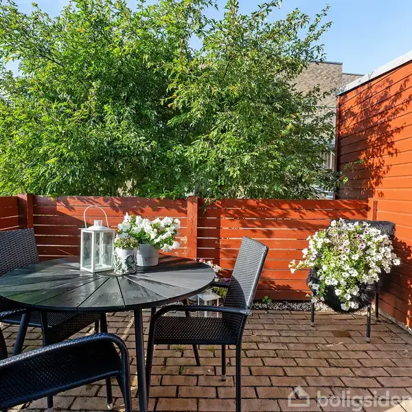 Et rundt bord med lanterner og stole står på en flisebelagt terrasse. Omgivet af rød træhegn, blomsterkrukker, og en grøn busk i baggrunden.