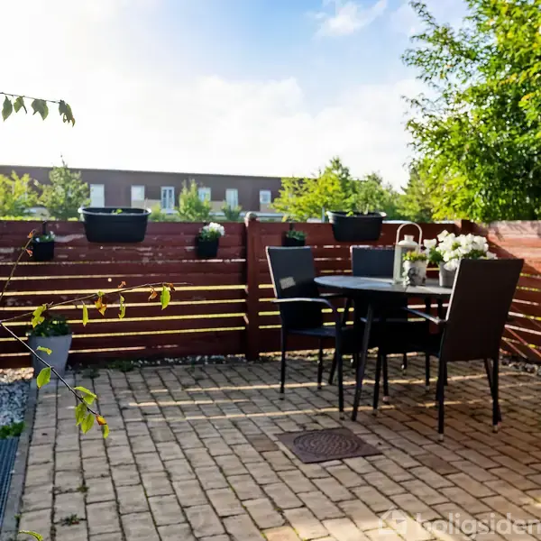 Terrasse med bord og fire stole omgivet af træhegn. Planter i krukker pryder omgivelserne. Solbeskinnet, med tæt bevoksning og en bygning i baggrunden.