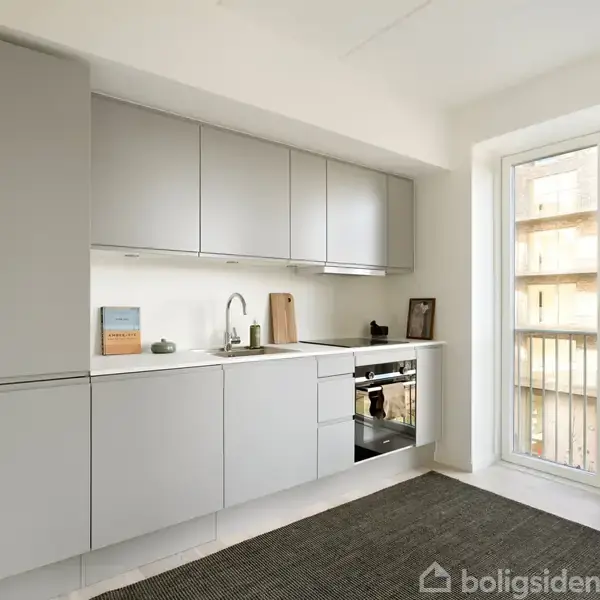 Køkken med grå skabe, ovn og vask, placeret langs en væg. En stor glasdør fører ud til en balkon. En mørk løber ligger på gulvet.