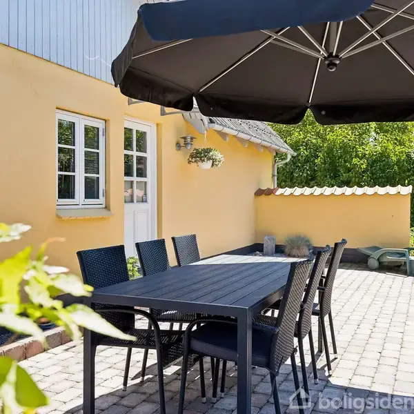Bord med sorte stole står under en stor parasol på en flisebelagt terrasse ved en gul husmur med vinduer og en dør; omgivet af grøn vegetation.