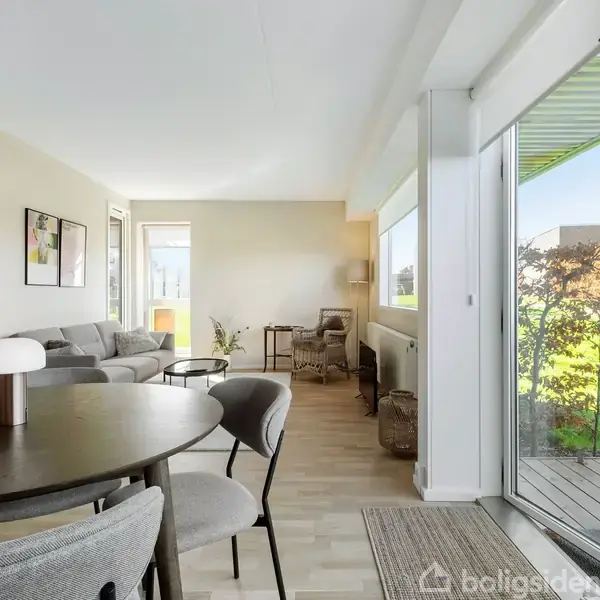 Rundt bord med stole står i fokus, placeret i en lys stue. Sofaer og skrivebord ses i baggrunden, store vinduer åbner op til en grøn have.