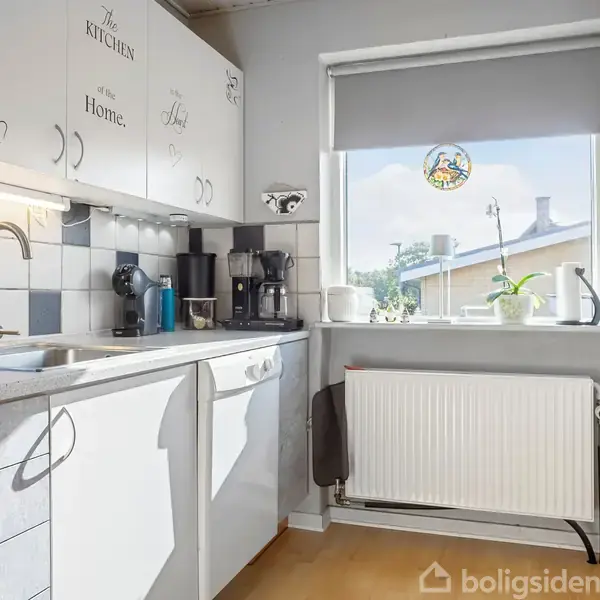 Et køkken med hvide skabe med teksten “The kitchen is the heart of the home.” En vask og kaffemaskine på bordet. Et vindue med udsigt til tage og planter i vindueskarmen.
