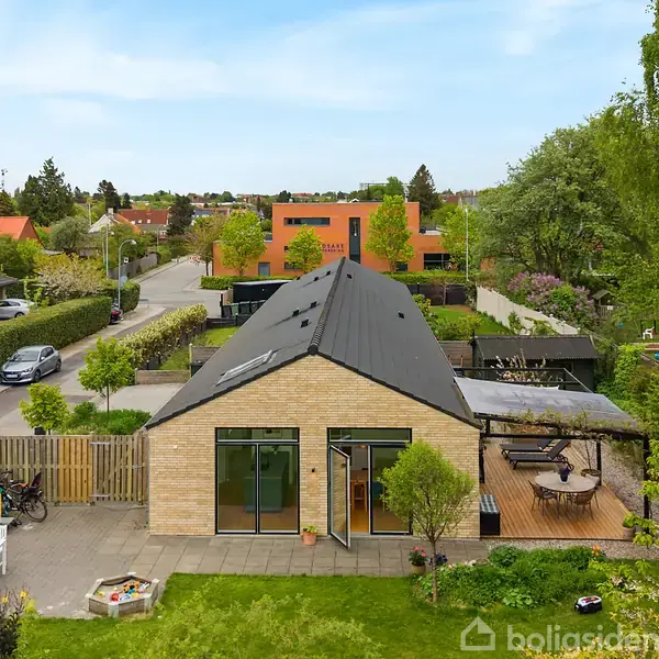 Et moderne, enkelt hus med fladt tag står på en grøn, velholdt grund. En flisebelagt terrasse er foran huset, som er omgivet af andre huse og træer i et kvarter.