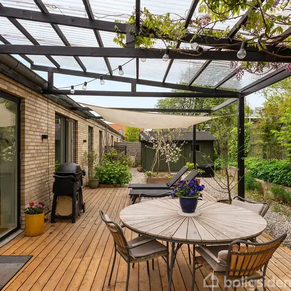 En træterrasse med et rundt bord og stole. Bordet har en blomsterpotte. En grill står tæt ved huset. En pergola med slyngplanter og hængende lys omfatter området, omgivet af grønne planter og et skur i baggrunden.