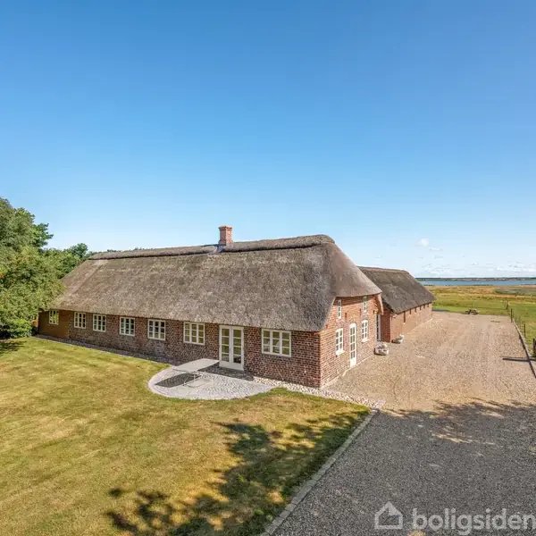 Stråtækt hus står stille på en åben mark ved en grusvej, omgivet af græsplæne og enkelte træer. Fjern udsigt over horisonten under en klar blå himmel.