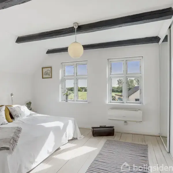 Soveværelse med dobbeltseng dækket af tæpper og puder. To vinduer slipper lys ind, spejler på garderobedøre reflekterer rummet. Hvidmalede bjælker og træplanker skaber en lys, luftig atmosfære.