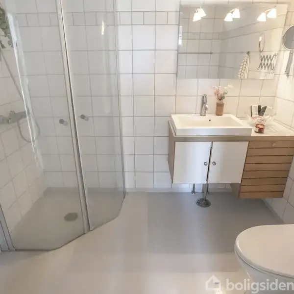 Et moderne badeværelse med en brusekabine til venstre. Vasken er monteret på et skab med træfinér, omgivet af hvide fliser, spejl og belysning. En toiletkumme ses i forgrunden.