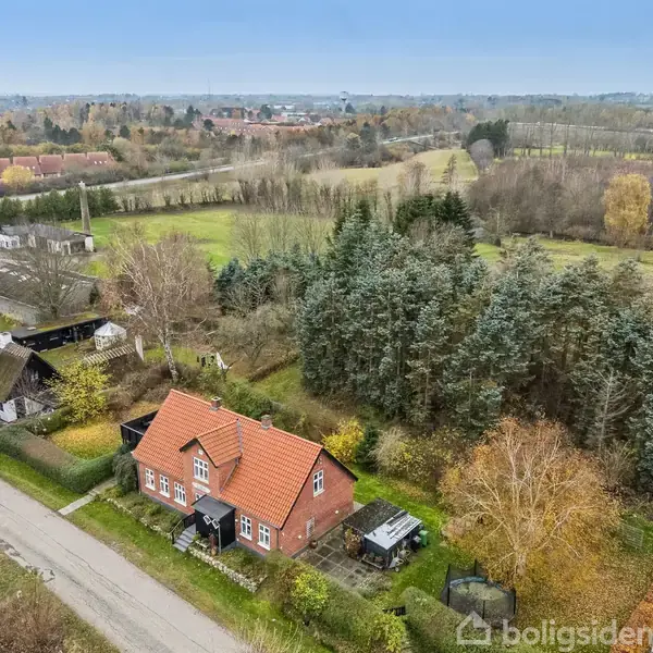 Rødt hus med rødt tag står stille langs en landevej. Omkring huset er der grønne skove og åbne marker. I baggrunden ses flere bygninger og åbne landskaber.