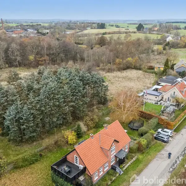 Hus med rødt tag ligger ved en vej omgivet af skove og marker. Flere biler parkeret ved huset. Foran huset er der træer, og små huse ligger langs vejen.