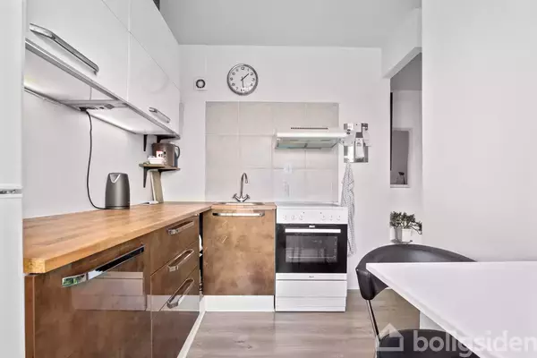 Køkken med hvide skabe og træbordplade, komfur i midten, ur på væggen. Lille spisebord i forgrunden, flisevæg bagved. Ingen tekst til stede.