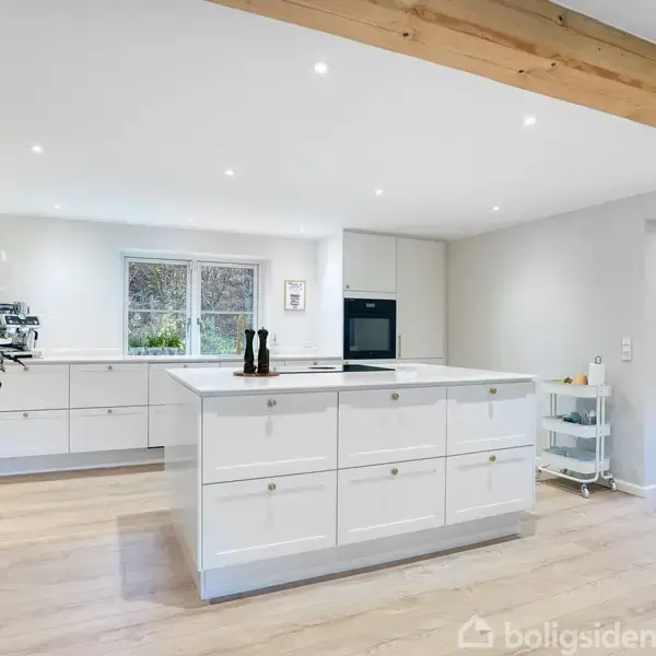 Køkkenø står centralt i rummeligt, lyst køkken med hvide skabe og trægulv. En plante står til venstre, og et stort vindue oplyser rummet.