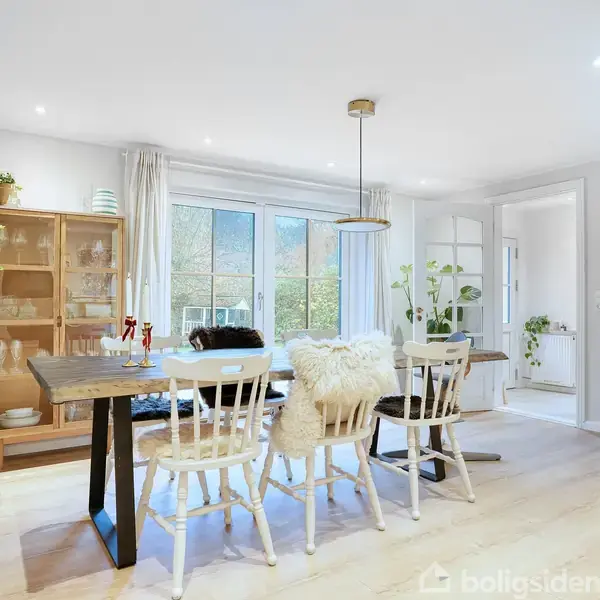 Et spisebord med seks stole er centralt placeret, omgivet af lyse vægge og store vinduer. En brændeovn brænder i hjørnet, mens planter og en reol fremhæver rummet.