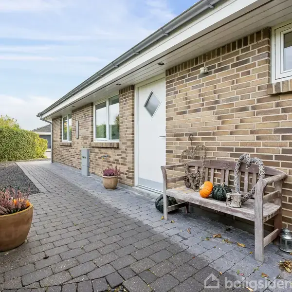 En murstensbygning med en hvid dør står i en flisebelagt havegang. Bænken ved siden af døren er dekoreret med en græskar og lanterner. Hækken omgiver området.