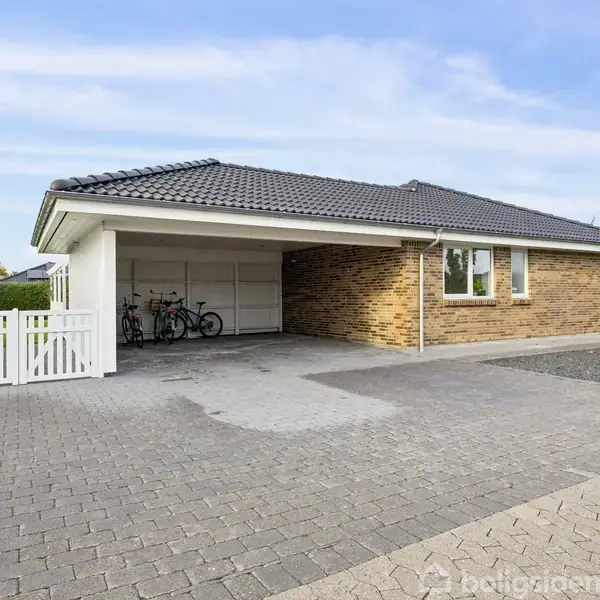 En murstensvilla med fladt tag står stille ved en bred indkørsel. Cykler er parkeret i en carport. Et hvidt stakit og grønne buske omgiver grunden under en blå himmel.