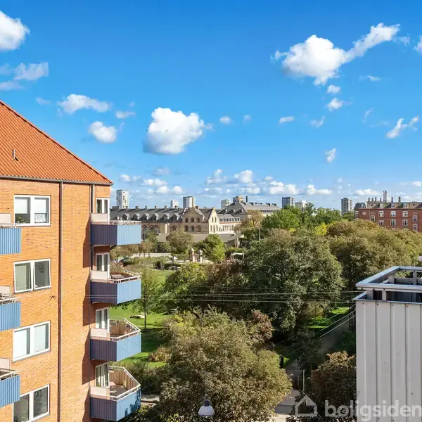 Røde murstensbygninger med blå altaner står i solskin. Udsigten viser en grøn park med træer. Baggrunden inkluderer flere bygninger under en klar blå himmel med spredte hvide skyer.