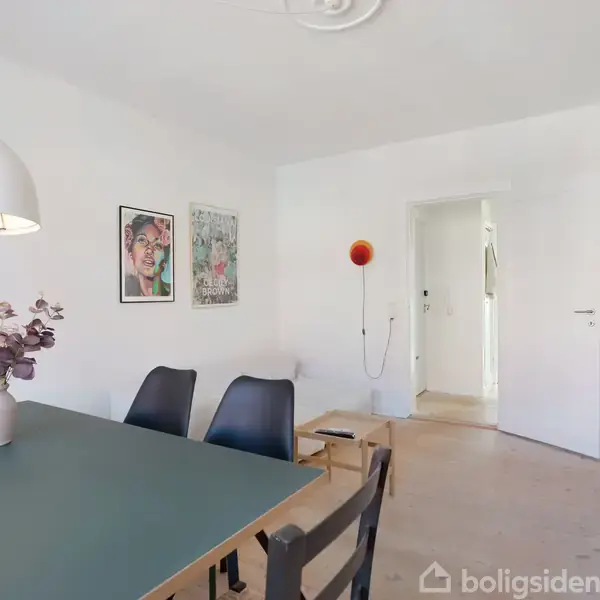Et spisebord med sorte stole står i et lyst rum. En hvid lampe hænger over bordet, og blomster i en vase pynter. Væggen har farverige plakater.