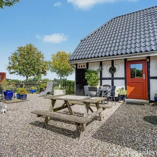 Et træbord og bænke står på en gårdsplads med grus, omgivet af røde bygninger og grønne træer. Der er også blomsterkrukker og et lille hegn.