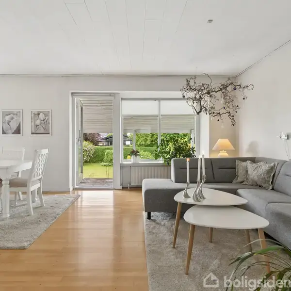 Stue med gråt hjørnesofaarrangement og sofabord, oplyst af en stålampe. Ved siden af står et spisebord med seks stole. Store vinduer fører ud til en have med grønne buske.