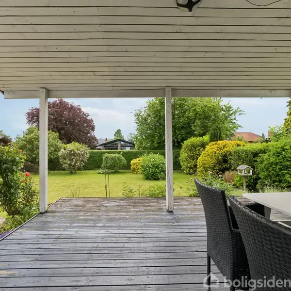 Overdækket terrasse med sorte kurvemøbler, der står stille. Den leder ud til en frodig have med grønne buske og træer i en solrig, klar dag.