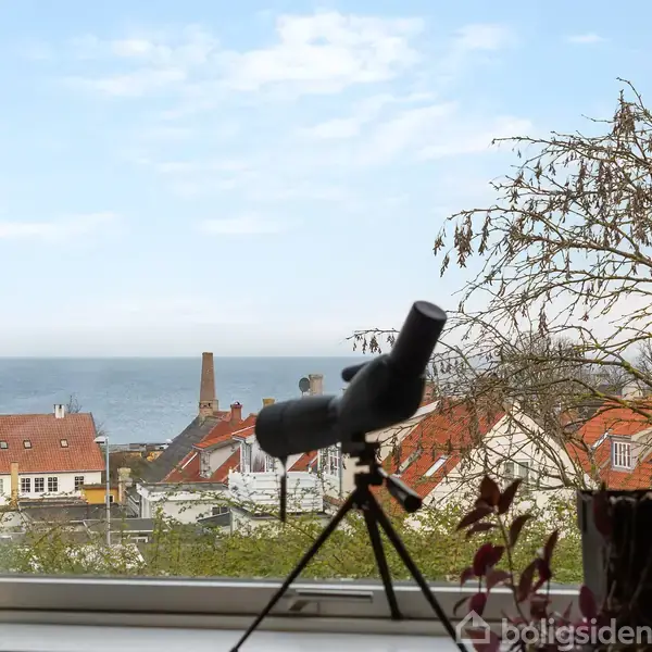 Kikkert står på vindueskarm og peger mod havudsigt, med røde hustage og træer i forgrunden.