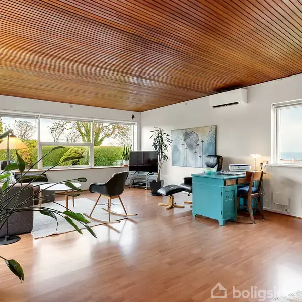 Stue med træloft og store vinduer, der giver udsigt til en have. En sofa og et fjernsyn står ved væggen. Et skrivebord med en blå stol er ved vinduet.