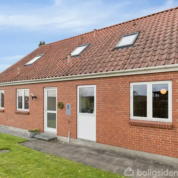 Et rødt murstenshus med rødt tegltag har flere vinduer og en indgangsdør. Huset står på en flisebelagt terrasse omgivet af en græsplæne under en blå himmel.