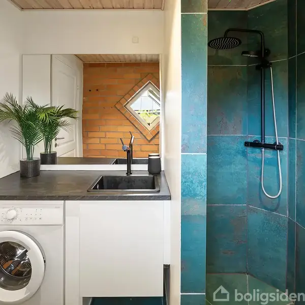 Vaskemaskine under en hvid bordplade med indbygget vask, omgivet af grønne fliser i bruseområdet. Et spejl og en plante står på bordpladen. Mørkt gulv og lys træloft.