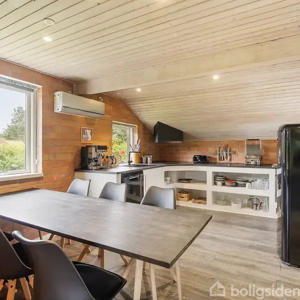 Et lyst køkken med spisebord og fire stole foran. Køkkenbordet har apparater, som en kaffemaskine, mens en sort køleskab står til højre. Rummets vægge er af mursten.