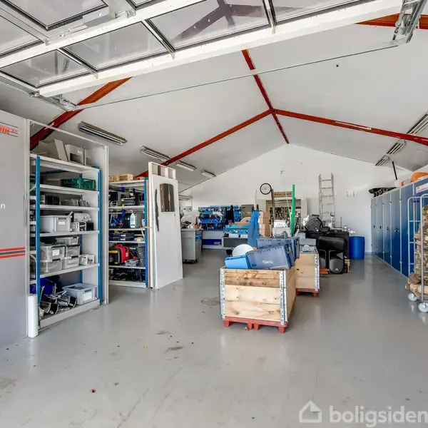 Værksted med hylder fyldt med værktøj og materialer; en central arbejdsbænk omgivet af opbevaringsskabe og værktøjskasser; rummet er lyst med et højt loft og lange lysrør. Tekst: "Eme dispenser, Prægeværktøjer, SATO Printer, NOVEXX Printer, Label MAX,".