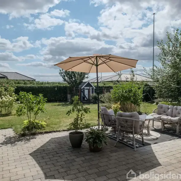 Terrasse med parasol og møbler som stående under i loungemøblering. Omgivet af frodig have med forskellige træer og buske. Blå himmel med skyer.