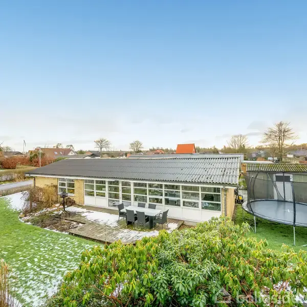 En lav bygning med store vinduer står i en have med sporadisk sne og grønne buske. Ved siden af er en trampolin. Husene og træerne omgiver området under en klar blå himmel.