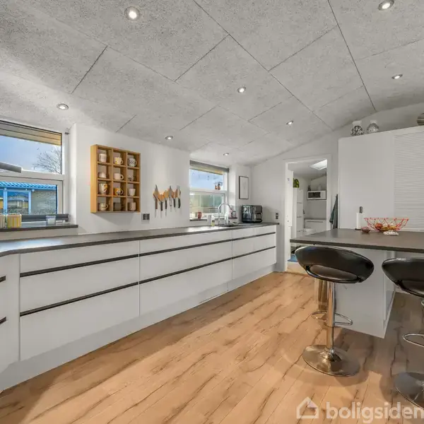 Et moderne køkken med hvide skabe og to sorte barstole står ved en morgenmadsbar. Store vinduer giver naturligt lys, og der er køkkenredskaber på væggen. Gulvet er træbelagt.