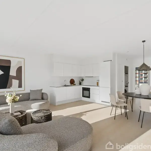 Stue med moderne møbler; en rund, grå sofa og sofaborde til venstre; spisebord og stole til højre. Baggrunden viser et lyst køkken med indbyggede skabe og en ovn.