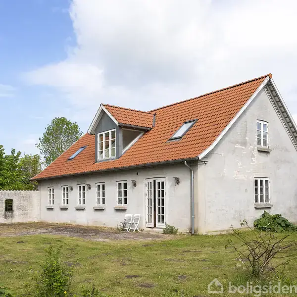 Hus med rødt tegltag og hvid facade står stille i en have med græsplæne og få buske. Stueetagen har flere vinduer, og baggrunden har tæt beplantning.