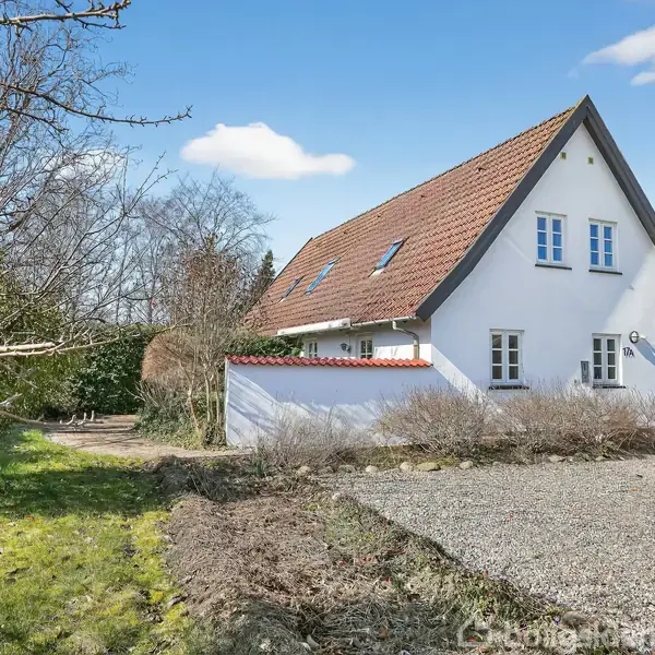 Et hvidt hus med rødt tegltag står i en have med grussti. Buske omgiver huset, og der er træer i baggrunden under en blå himmel med få skyer.