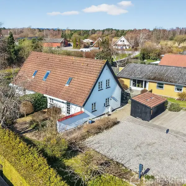 Et rummeligt, hvidt hus med rødbrunt tegltag ligger ved siden af en gul bygning med sort tag. Huset har en indkørsel omgivet af træer og grønne buske i et landligt område.