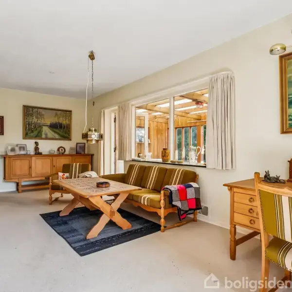 Stue med træmøbler inkluderer en sofa og sofabord, placeret på tæppe foran vindue. Væggene er dekoreret med billeder, og der er et vitrineskab i baggrunden.