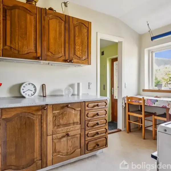 Et køkken med træskabe og en hvid bordplade indeholder en hvid analog vækkeur. Bordet ved et vindue er dækket med en farverig dug og omgivet af stole.