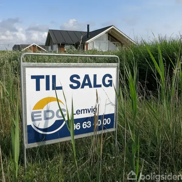 Et skilt står i græsset med teksten "TIL SALG, EDC Lemvig, 96 63 00 00". I baggrunden ses et hus omgivet af lave buske og blå himmel.