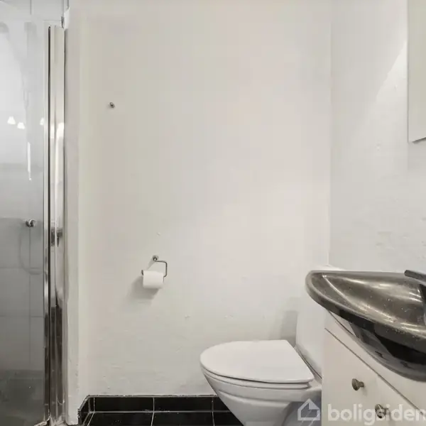 Toilet står stille i et lyst badeværelse med en hvid væg. Ved siden af er der et håndvask med et spejl over. En brusekabine er til venstre.