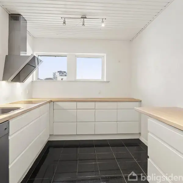 Et lyst køkken med hvide skabe, sorte fliser på gulvet og træbordplader. Der er en indbygget ovn, opvaskemaskine og vask. Et stort vindue giver naturligt lys.