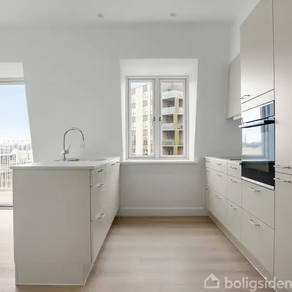 Et lyst, moderne køkken med hvide skabe og skuffer. En ø med indbygget vask står centralt. Store vinduer til venstre og midten giver udsigt til altan og lejlighedsbygning.