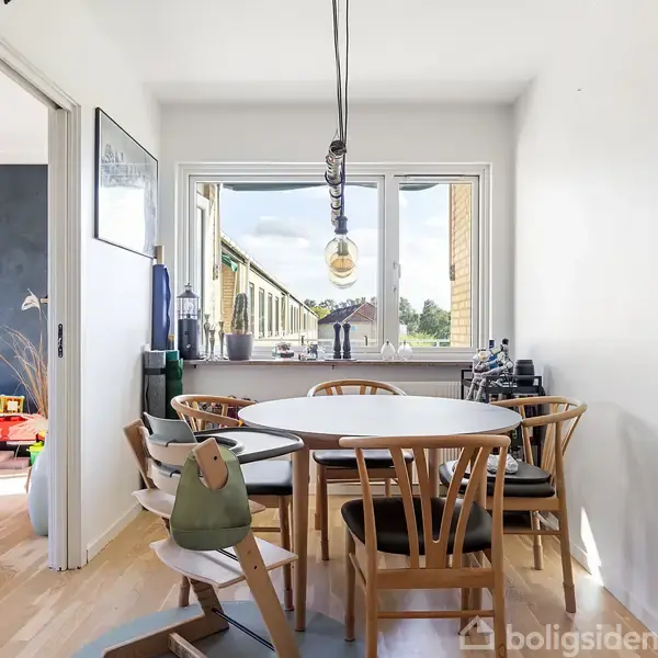 Et rundt spisebord med seks stole står i et lyst rum. Et stort vindue bagved viser udsigt til en altan. Til venstre er der en åben dør til et legetøjfyldt værelse.
