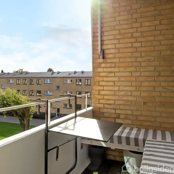 Balkon med udsigt over en græsplæne og flere boligbygninger. Der er et lille bord og en bænk med stribepuder langs murstensvæggen. Sollys skinner ind fra venstre.
