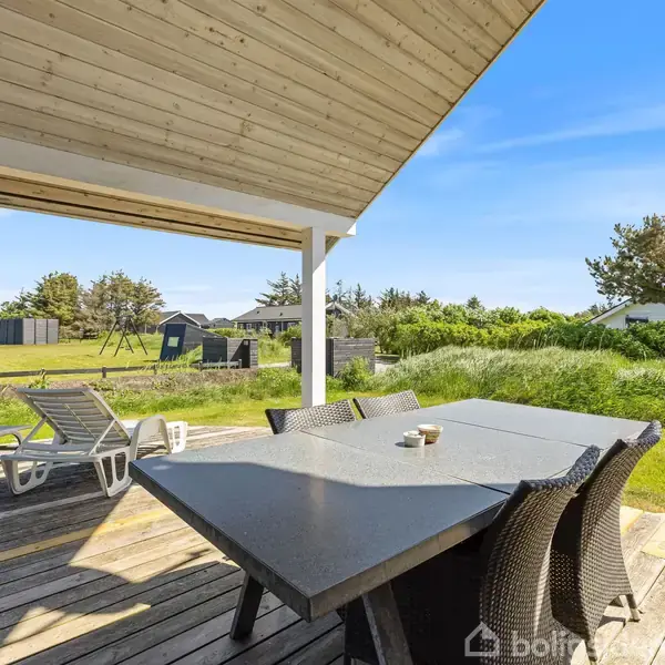 Terrassebord med stole står på en træterrasse. Ved siden af er der liggestole. I baggrunden ses en grøn græsplæne og spredte træer under en klar blå himmel.