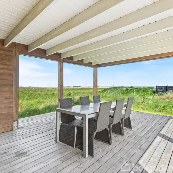 Spisebord med seks stole står på en overdækket terrasse i træ. Til venstre ses en grill. I baggrunden er der udsigt over en eng og blå himmel.